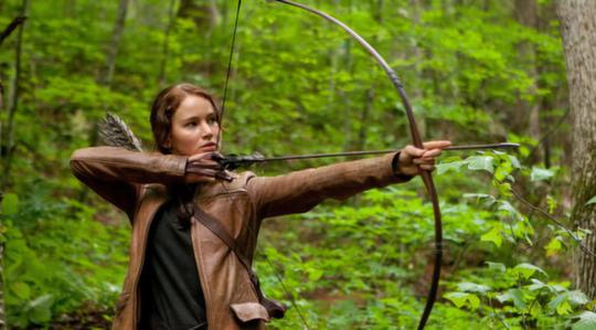 Katniss aiming with her bow in the forest outside district 12